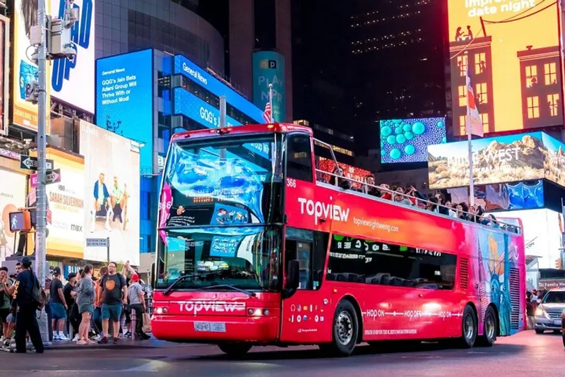 Open-top double-decker on a NYC bus tour at night with Times Square lit up behind the upper deck